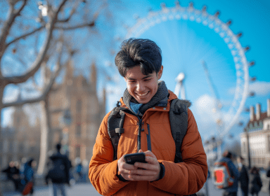 A cheerful young person is standing outdoors, holding and looking at a smartphone with a smile. They're wearing a winter jacket and backpack, suggesting they might be traveling or commuting. The scene is set against a clear blue sky.