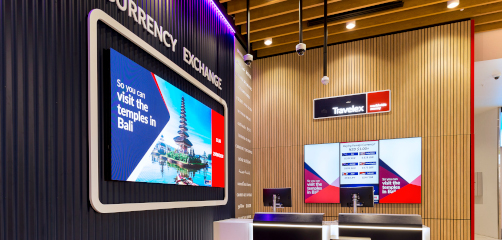 Interior of a modern currency exchange store with a vibrant digital display promoting travel to Bali, complemented by a sleek black counter and informative screens showing exchange rates. The Travelex logo is prominently displayed above.