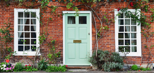 A quaint red brick house front with a central light green door flanked by two white-framed windows, adorned with climbing vines and surrounded by a small flowerbed featuring colorful blooms.