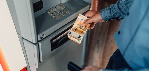 A person withdrawing currency from an ATM, holding a crisp fifty euro note. The focus is on the banknote and the ATM keypad, highlighting the convenience of cash access.